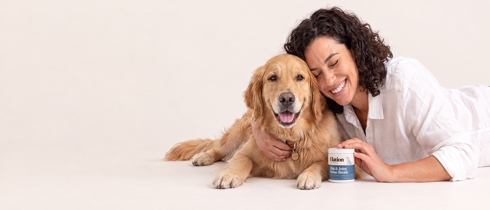 A laughing woman with her eyes closed hugs her smiling golden retriever on a white studio floor, her fingertips on a small jar of Elation Hip & Joint Bone Broth.