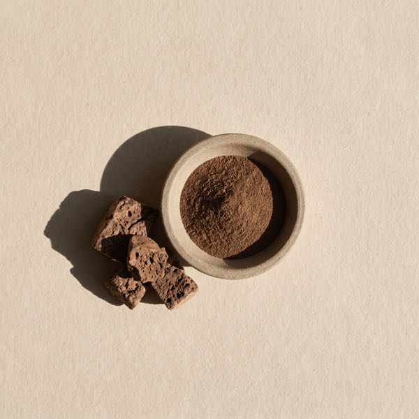 Pieces of dried beef liver beside a small stoneware dish of dark-brown liver powder, on a stone-colored background.